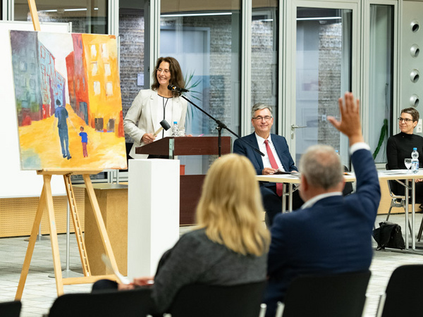 24.09.2021 - Kunstversteigerung mit Bürgermeisterin Andrea Schwarz Eine Frau steht lächelnd hinter einem Rednerpult und spricht zu einem Publikum. Im Hintergrund ist ein buntes Gemälde mit einer Szene von Menschen zu sehen. Einige Zuhörer sitzen im Vordergrund, einer hat die Hand erhoben, als Zeichen, dass er eine Frage stellen möchte.