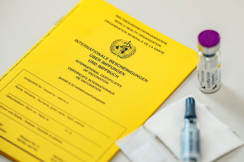 Ein gelbes Impfheft mit der Aufschrift "Internationale Bescheinigungen über Impfungen" liegt auf einem Tisch. Daneben befinden sich eine Spritze und eine Ampulle mit Impfstoff auf einem weißen Tuch. (Text KI generiert)