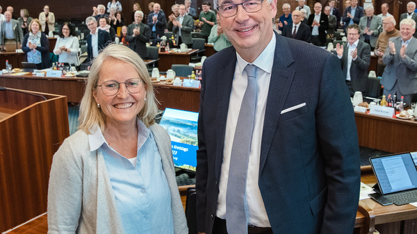 Due persone sorridenti sono al centro dell'immagine, con una folla di immagini sfocate che applaude sullo sfondo. L'ambientazione è un'aula, probabilmente un incontro ufficiale o una conferenza. Le due persone sembrano professionisti pronti a scambiare un momento di celebrazione.