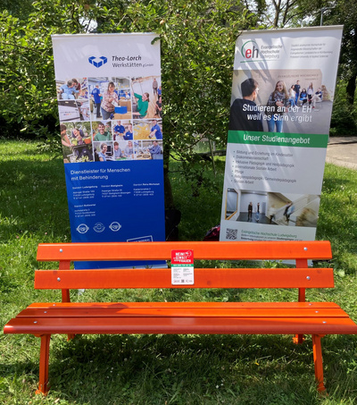 L'immagine mostra una panchina arancione posizionata in uno spazio verde. Sullo sfondo, ci sono due banner informativi, uno con il logo di Theo-Lorch e l'altro riguardante opportunità di studio. Gli elementi sono disposti in un contesto all'aperto.