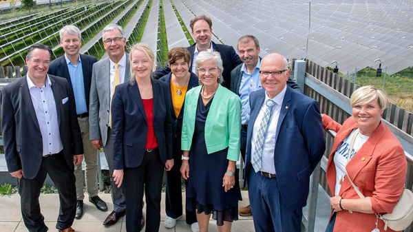 Eine Gruppe von zehn Personen steht zusammen auf einem Platz, im Hintergrund sind Solarpanels und ein Wasserturm sichtbar. Die Gruppe besteht aus Männern und Frauen unterschiedlichen Alters, die lächeln und in formeller Kleidung auftreten.