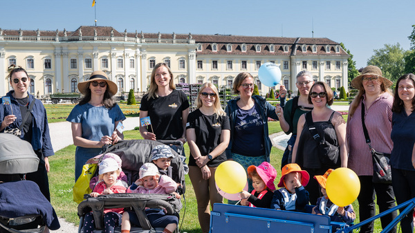 Eine Gruppe von Frauen und Kindern steht in einem Park vor einem beeindruckenden Gebäude. Einige Frauen tragen Sonnenhüte, und im Vordergrund sind Kinder in Kinderwagen und ein Wagen mit Luftballons zu sehen. Der Himmel ist klar und blau.