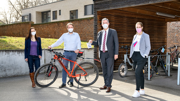 Four individuals stand outdoors near a bicycle. They are smiling and wearing masks. One person holds a bike, while another holds a piece of paper, possibly a document related to the bike. The setting appears to be a sunny day in a residential area.