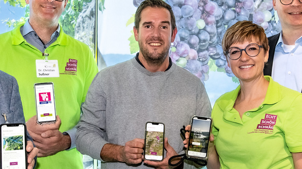 Fünf Personen stehen lächelnd vor einer Wand mit Weintraubenmotiven. Sie halten jeweils ein Smartphone in der Hand, auf deren Bildschirmen verschiedene Apps zu sehen sind. Alle tragen grüne T-Shirts oder Hemden.