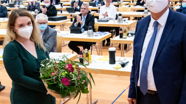 A woman in a green dress receives a bouquet of flowers from a man in a suit, both wearing masks. They stand in a large hall filled with people seated at tables, applauding. The atmosphere is celebratory, likely during an official event or ceremony.