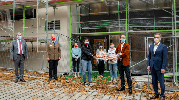 A group engaged in a construction-related event stands outside a building under renovation. Some individuals wear masks and hold a large tray of bread, likely for a ceremonial occasion. The scene conveys a sense of community and celebration amidst ongoing construction.