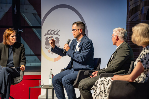 Eine Podiumsdiskussion mit vier Personen: Ein Mann in Anzug hält ein Mikrofon und zeigt einen Gegenstand. Zwei weitere Männer und eine Frau hören aufmerksam zu. Im Hintergrund ist ein Banner mit einem stilisierten Wappen. Die Szene vermittelt einen professionellen und interaktiven Austausch.