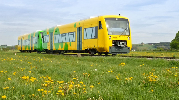 A bright yellow train with a green design travels along a railway amidst a field of dandelions. The landscape features lush grass and a tree in the background, suggesting a rural setting under a partly cloudy sky.