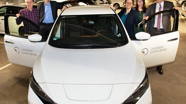Five individuals stand around a white Nissan electric vehicle in a parking garage. The car has its front doors open. The people appear to be engaged in a discussion about the vehicle, representing a moment of collaboration or a launch event relating to electric transportation.