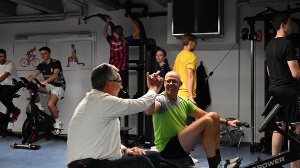 In einem Fitnessstudio trainieren mehrere Personen an verschiedenem Equipment. Zwei Männer sitzen auf Rudergeräten, während ein anderer Mann ihnen zuschaut und sie anfeuert. Im Hintergrund sind weitere Personen mit einer Kettlebell und auf Fahrradergometern zu sehen.
