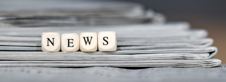 Header Bild: Eine Ansammlung von Zeitungen mit dem Wort "NEWS", dargestellt durch Holzbuchstaben, das zentral auf einem Stapel liegt. Die Zeitungen sind in Grautönen gehalten und geben einen Eindruck von aktuellen Informationen und Nachrichten.