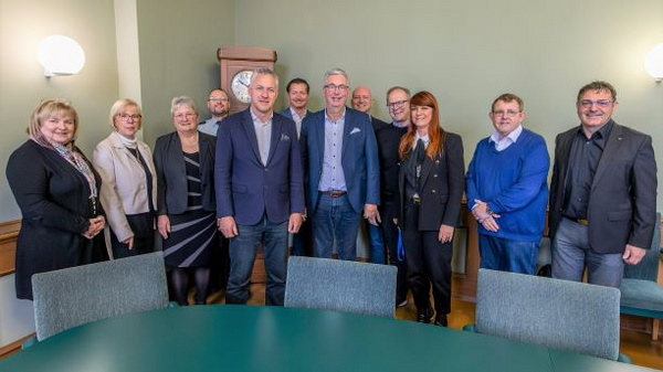 Gruppe von 12 Personen in einem Raum mit einem großen Tisch. Sie stehen zusammen, lächeln und tragen formelle Kleidung. Im Hintergrund ist eine Uhr an der Wand zu sehen. Die Personen scheinen an einem offiziellen Anlass oder Treffen teilzunehmen.