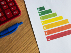 Das Bild zeigt ein Blatt Papier mit einer farbigen Skala von A bis G, die in grün, gelb und rot verlaufen ist. Daneben liegen ein roter Taschenrechner und ein blauer Stift auf einem Holztisch.