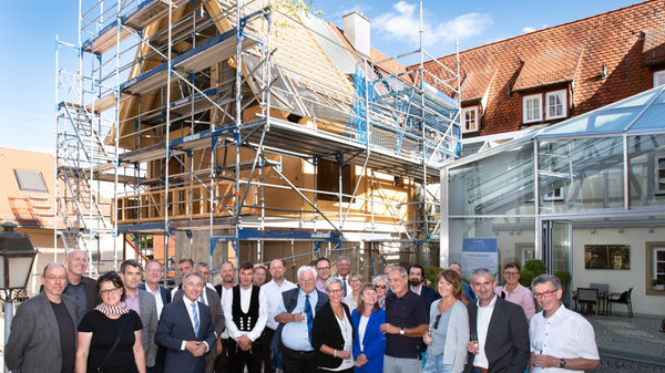 A group of about 25 people, including men and women, stands in front of a partially constructed building surrounded by scaffolding. They appear to be celebrating or marking an event, with a clear blue sky in the background.
