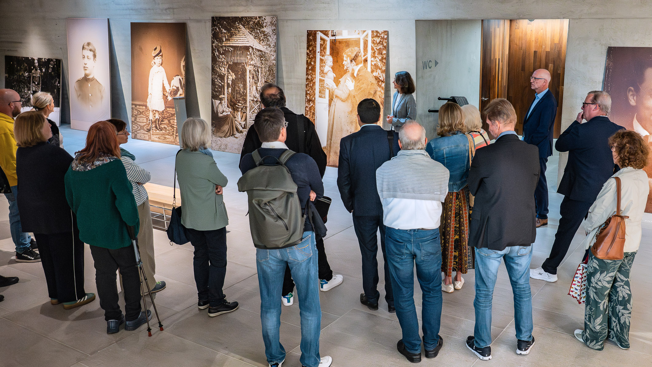 Eine Gruppe von Menschen steht in einem Ausstellungsraum vor großen Fotografien an der Wand. Eine Person spricht zu den Anwesenden. Das Ambiente wirkt informell und fördert den Austausch über die ausgestellten Werke.