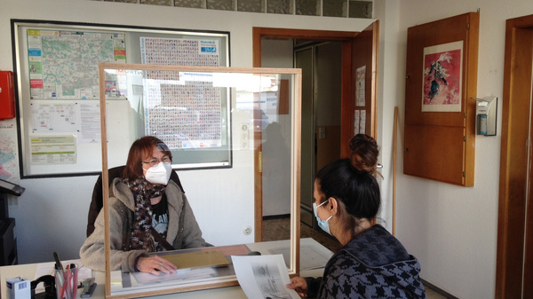 A person wearing a mask sits behind a protective acrylic barrier at a reception desk, assisting another masked individual who is seated and holding documents. The background includes a map on the wall and doors.