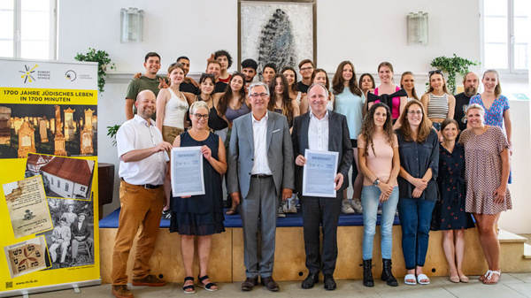 Un gruppo di circa trenta persone sorride insieme, alcune tenendo dei documenti. Al centro, due uomini in abiti formali reggono un documento, circondati da giovani adulti. Sullo sfondo, un banner che celebra 1700 anni di vita ebraica è visibile.