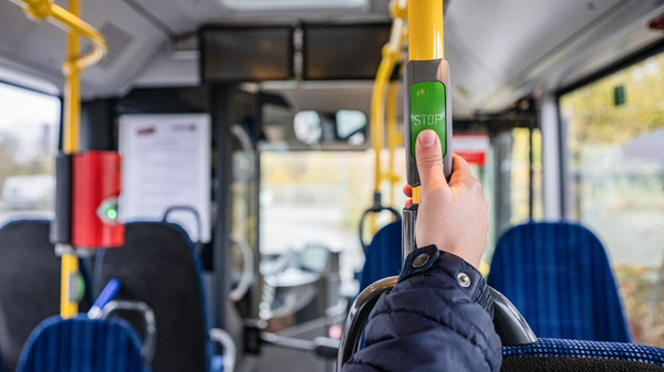Eine Hand hält eine Haltestange in einem Bus. An der Haltestange befindet sich ein grüner Knopf mit der Aufschrift „STOP“. Im Hintergrund sind Sitzplätze und die Fahrerkabine sichtbar.