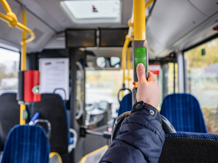 Eine Hand hält eine Haltestange in einem Bus. An der Haltestange befindet sich ein grüner Knopf mit der Aufschrift „STOP“. Im Hintergrund sind Sitzplätze und die Fahrerkabine sichtbar.