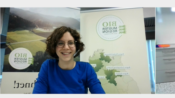 Ein Online-Workshop zur Zubereitung von Speisen. Eine Frau in einem blauen Oberteil lächelt von der linken Seite des Bildes, während eine andere Frau in einer weißen Schürze auf der rechten Seite eine Schüssel mit Teig hält. Hinter ihnen sind verschiedene Küchenutensilien und ein Banner sichtbar.