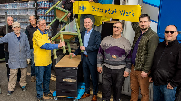 Un gruppo di dodici persone, alcuni in abiti da lavoro e altri in abbigliamento casual, posa insieme davanti a un camion. Al centro, un uomo in maglietta gialla tiene un cartello con la scritta "Gustav-Adolf-Werk". L'atmosfera è di collaborazione e abbraccio comunitario.