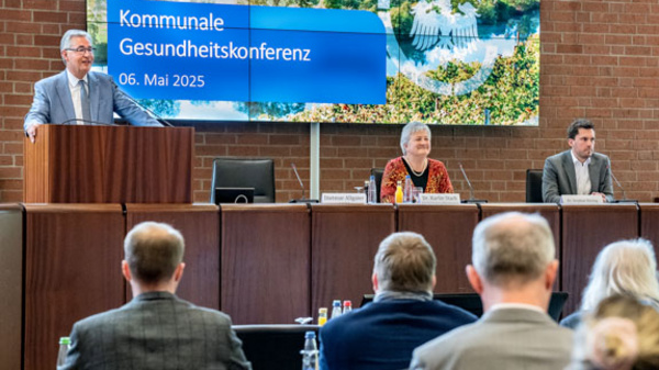 Bildbeschreibung: Eine Person steht am Rednerpult auf einer Konferenz zum Thema Gesundheit. Auf einem großen Bildschirm im Hintergrund steht „Kommunale Gesundheitskonferenz, 06. Mai 2025“. Die Zuschauer im Raum hören aufmerksam zu.
