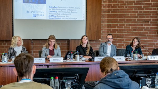 A panel of five speakers sits at a conference table during a press event. They appear to be discussing subjects related to education and finance. A large screen displays information above them. Attendees are seated in the foreground, focusing on the speakers.