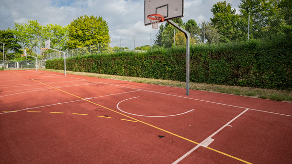 Basketballfeld mit einer Korbanlage. Der Platz ist umgeben von hohen Büschen und Bäumen, der Boden ist rot mit Markierungen für das Spiel. Himmel mit einigen Wolken ist sichtbar. (Text KI generiert)