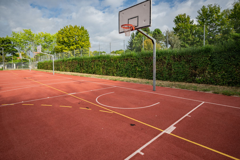 Basketballfeld mit einer Korbanlage. Der Platz ist umgeben von hohen Büschen und Bäumen, der Boden ist rot mit Markierungen für das Spiel. Himmel mit einigen Wolken ist sichtbar. (Text KI generiert)