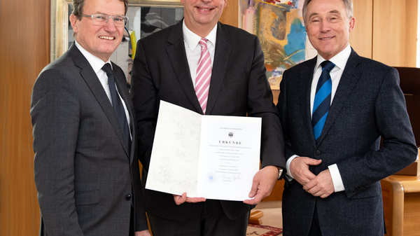 Three men stand in an office setting, smiling and posing together. The central figure holds a formal document, while the others stand beside him, looking pleased. The background features wooden panels and decorative artwork, suggesting a professional atmosphere.