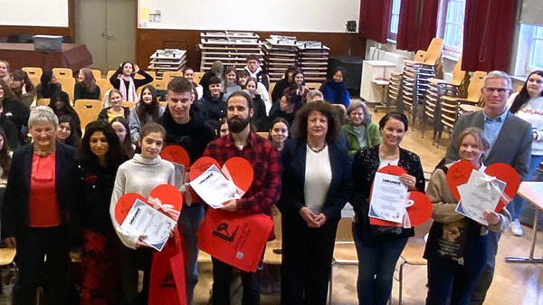 Eine Gruppe von Menschen steht in einem Raum, umgeben von Stühlen und Tischen. Viele halten rote Herzen und Zertifikate in der Hand. Die Anwesenden lächeln, während sie für ein Gruppenfoto posieren. Der Raum hat große Fenster und eine freundliche Atmosphäre.