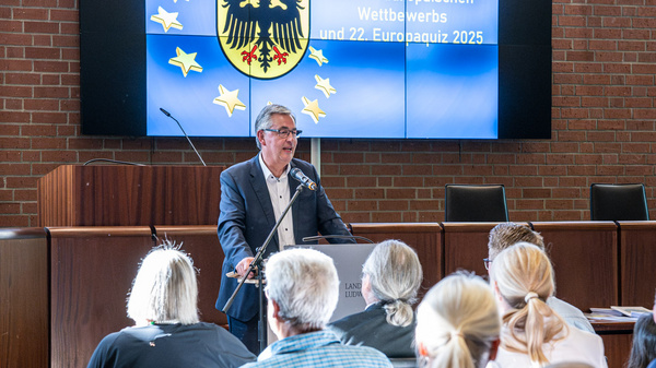 Eine Person steht an einem Rednerpult vor einem Bildschirm, der mit „Preisverleihung des 72. Europäischen Wettbewerbs“ beschriftet ist. Im Hintergrund sitzen Zuhörer, die aufmerksam zuhören. Die Atmosphäre ist feierlich und informativ.