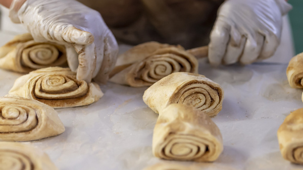 Eine Person mit Handschuhen formt Zimtschnecken aus Teig. Die Zimtschnecken sind spiralförmig auf einem Backblech angeordnet. Der Hintergrund ist unscharf, wobei der Fokus auf dem Teig und der Herstellung liegt.