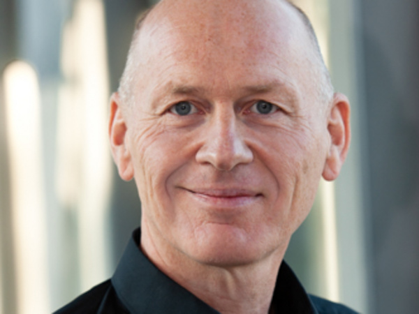 A middle-aged man with a shaved head and a slight smile is wearing a black shirt. The background features soft, blurred light, suggesting an indoor setting. He appears confident and approachable.