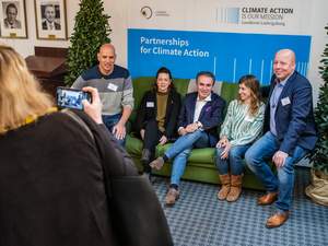 Eine Gruppe von fünf Personen posiert auf einem Sofa für ein Foto. Im Hintergrund steht eine Wand mit dem Schriftzug "Climate Action is our Mission" und "Partnerships for Climate Action". Grüne Pflanzen sind ebenfalls sichtbar.
