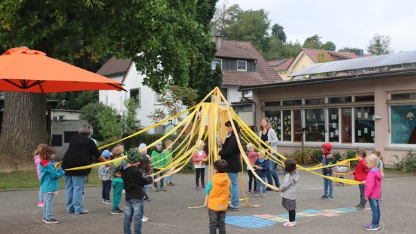 Des enfants et des adultes participent à une activité en plein air, entourés de rubans colorés qui forment une structure centrale. Les enfants sont joyeux, vêtus de vestes et de bonnets, tandis que les adultes les encouragent dans cette activité ludique.