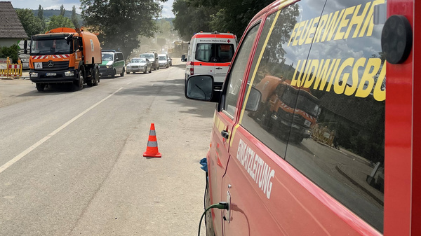 Une camionnette de pompiers rouge est garée sur le bord d'une route, avec des véhicules en attente derrière elle. Des cônes de sécurité sont disposés sur la chaussée. Le ciel est partiellement nuageux, et la végétation est présente au bord de la route.