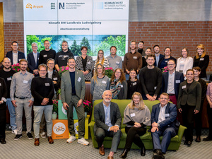 Eine Gruppe von Menschen steht lächelnd vor einem Banner mit dem Text „Klimafit im Landkreis Ludwigslust-Parchim“ und weiteren Informationen. Die Gruppe besteht aus Männern und Frauen verschiedener Altersgruppen, die alle positive Energien ausstrahlen. Im Vordergrund sind einige Personen sitzend.