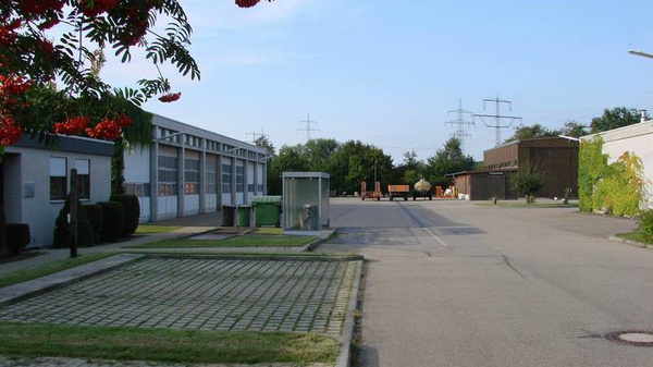 Une vue d'un espace urbain avec des bâtiments industriels sur la gauche et une allée pavée à droite. Un abribus se trouve le long de la route, bordé de verdure. Le ciel est dégagé et offre une lumière naturelle.