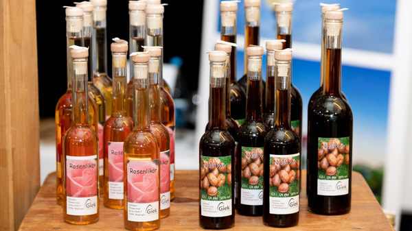 A wooden display features various bottles of syrup. There are two distinct types: clear, golden syrups labeled "Rosenliker" and dark syrups, each adorned with a label depicting nuts. The arrangement highlights multiple bottles, showcasing their elegant designs and colors.