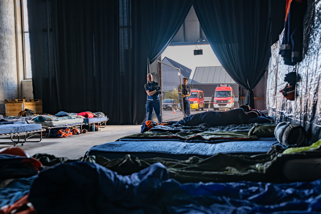 Im Inneren einer Halle sind Liegematten auf dem Boden ausgelegt. Zwei Personen stehen in der Nähe des Eingangs und schauen nach draußen, wo Feuerwehrfahrzeuge zu sehen sind. Die Atmosphäre wirkt ruhig und auf Unterstützung ausgerichtet.