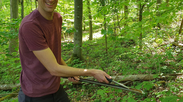 A smiling man stands in a lush green forest, holding a pair of pruning shears. He appears to be engaged in outdoor activities, surrounded by trees and foliage on the ground. Natural light filters through the leaves, creating a vibrant atmosphere.