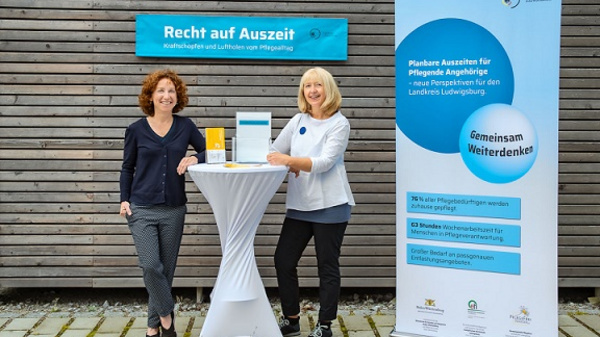 Deux femmes se tiennent à côté d'une table au centre, avec un dépliant et un support d'information. Une affiche à droite présente des informations sur le droit à des pauses pour les aidants. En arrière-plan, un panneau bleu indique "Recht auf Auszeit".