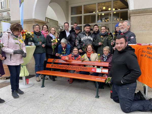 Un gruppo di persone si riunisce attorno a una panchina arancione con la scritta "#schweigenbrechen". Alcuni partecipanti tengono fiori, mentre altri sorridono. L'evento sembra essere una manifestazione per sensibilizzare contro la violenza. Il contesto è un ambiente urbano e ben illuminato.