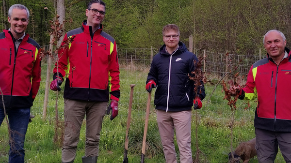 Vier Personen in roten Jacken stehen im Freien und halten Setzlinge in der Hand. Sie stehen auf einem grünen Feld, umgeben von Bäumen. Die Gruppe lächelt und posiert, bereit für eine Pflanzaktion. Ein Eimer und Werkzeuge liegen auf dem Boden.