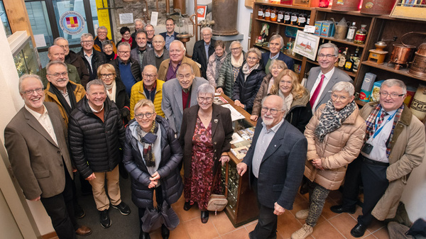 Eine Gruppe von 34 Personen versammelt sich in einem Raum, umgeben von Regalen mit verschiedenen Gegenständen. Die Mitglieder zeigen ein Lächeln, und einige halten Utensilien oder stehen nah beieinander. Die Atmosphäre wirkt freundlich und einladend.