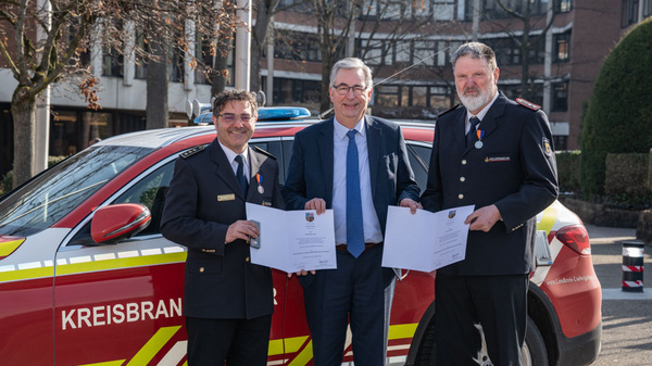 Drei Männer stehen vor einem Einsatzfahrzeug der Feuerwehr. Zwei von ihnen halten Urkunden in den Händen, während der dritte in der Mitte steht und lächelt. Alle tragen formelle Kleidung, und im Hintergrund sind Bäume und Gebäude zu sehen.