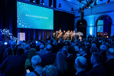 Eine Gruppe von Musikern tritt während einer festlichen Veranstaltung auf, die als Neujahrsempfang 2026 bezeichnet wird. Das Publikum sitzt gebannt im Vordergrund, während eine große Leinwand im Hintergrund das Event-Thema anzeigt. Die Atmosphäre ist festlich und formell.