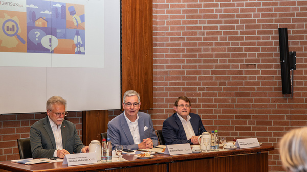 Drei Männer sitzen an einem Tisch bei einer Veranstaltung. Im Hintergrund ist eine Präsentation mit Grafiken und Diagrammen zu sehen. Die Männer scheinen über ein wichtiges Thema zu sprechen, während auf dem Tisch Getränke und Notizen liegen.