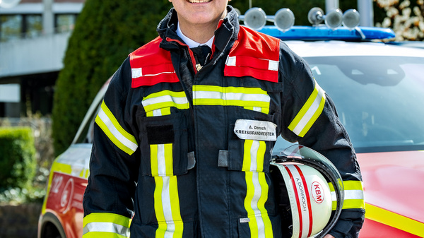 Ein Feuerwehrmann steht lächelnd vor einem Einsatzfahrzeug. Er trägt eine auffällige Feuerwehrjacke mit reflektierenden Streifen und hält einen Helm in der Hand. Im Hintergrund sind Bürobauten und grüne Bäume sichtbar.